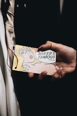 A hand is holding a vintage-style tag featuring a pop art illustration of a blonde woman alongside text that reads 'Naked & Famous Denim'. The background is dark, and the focus is on the tag.