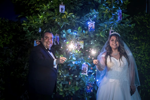 A bride and groom stand in front of a lush green backdrop adorned with string lights and several small photos. They are both holding sparklers, and the bride is dressed in an elegant white gown with a veil and tiara, while the groom is in a classic black tuxedo. Both are smiling and appear joyful.
