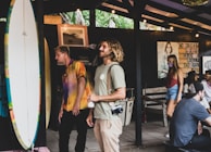 Several people are gathered in an open-air setting that seems to be a casual, relaxed environment. A surfboard is prominently displayed on the left side, while two men stand nearby, one with long hair and a camera around his neck. A woman in a red top and denim shorts walks by in the background, wearing a face mask. Decor includes a photograph, a quote on the wall, and ambient lighting.