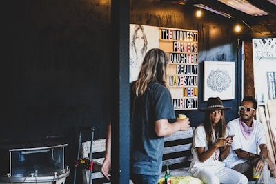 A casual setting with a group of three people. Two people are sitting, one wearing a wide-brimmed hat and the other in sunglasses with a bandana. A third person with long hair stands in front, holding a drink. A sign with colorful letters reads 'The Midwest Farmers Daughter Really Makes You Feel Alright.' The room has dim lighting with exposed bulbs, and there is artwork displayed on the dark walls.