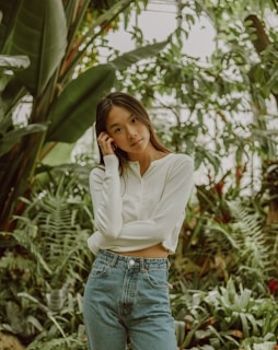 A person stands in a lush green environment, surrounded by various large leaves and plants. They are wearing a white long-sleeve shirt and blue jeans, with one hand touching their hair.