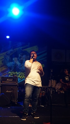 A performer is on stage holding a microphone, wearing a white shirt and jeans. Behind them, there is a backdrop with colorful graphics and a band setup with instruments like drums. Amplifiers are visible on stage, and stage lighting creates a vibrant atmosphere.