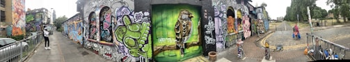 A street is lined with vibrant graffiti art covering the entire length of the building's walls. The artwork includes various colorful designs, characters, and intricate patterns. A bird painted on a door stands out in green hues. Two people are walking along the street, with urban scenery in the background.