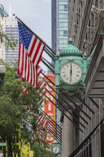 A large building facade featuring a green ornamental clock is adorned with multiple American flags. In the background, a prominent red and yellow sign spells out 'Chicago.' Trees and modern high-rise buildings are visible, highlighting an urban setting.