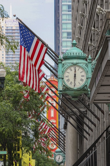 A large building facade featuring a green ornamental clock is adorned with multiple American flags. In the background, a prominent red and yellow sign spells out 'Chicago.' Trees and modern high-rise buildings are visible, highlighting an urban setting.