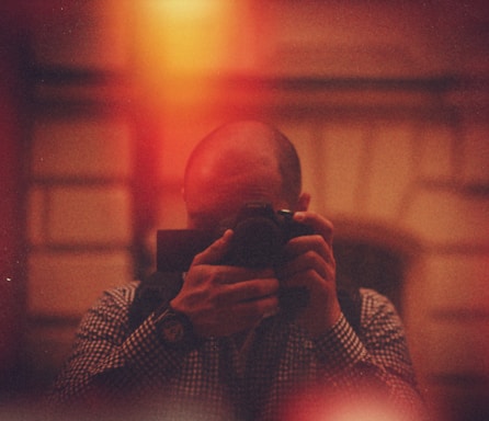 A person is holding a camera, capturing a photo in a dimly lit setting with a warm, reddish glow. The background appears blurred and abstract, adding to the overall atmospheric composition.