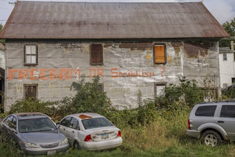 A worn-out, two-story building with boarded-up windows has graffiti reading 'Freedom or Socialism?' on its deteriorating gray facade. Overgrown bushes and three parked cars, including a silver sedan, a white car with a rusty roof, and a silver SUV, occupy the foreground. The roof is corrugated metal, and the surrounding area appears neglected.