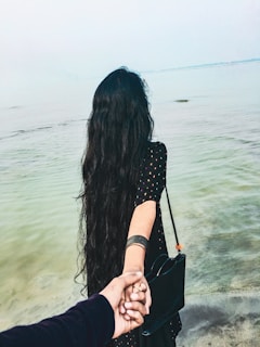 A person with long dark hair is standing near a body of water, holding another person's hand. The person in focus is wearing a black outfit and carrying a black bag. The scene conveys a sense of intimacy and connection.