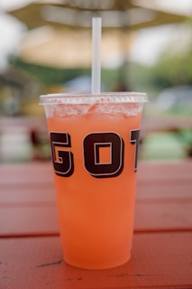 A clear plastic cup filled with an orange beverage, featuring a lid and a straw. The cup is situated on a wooden surface with a blurred outdoor background, suggesting a relaxed, outdoor setting.