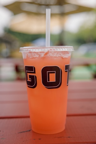 A clear plastic cup filled with an orange beverage, featuring a lid and a straw. The cup is situated on a wooden surface with a blurred outdoor background, suggesting a relaxed, outdoor setting.