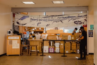 A small food stall is situated indoors and displays a banner featuring an illustration of a train with sharks. The stall has various stacked boxes and containers, and two individuals are present, wearing casual clothes and face masks. There are two wooden stools in front of the counter and a partition separating the workers from the public area.