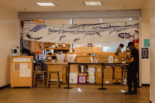 A small food stall is situated indoors and displays a banner featuring an illustration of a train with sharks. The stall has various stacked boxes and containers, and two individuals are present, wearing casual clothes and face masks. There are two wooden stools in front of the counter and a partition separating the workers from the public area.