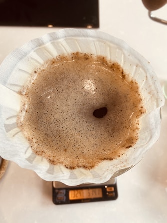 A close-up view of a coffee brewing process in a white paper filter, showing the bubbling surface of freshly brewed coffee. The filter rests on top of a digital scale, with readings partially visible below.