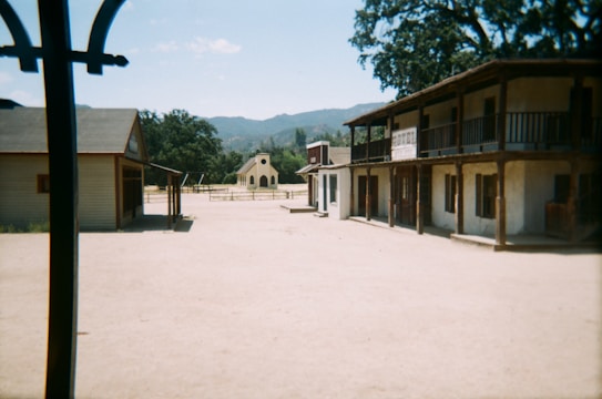 A deserted, old-fashioned Western-style town with dirt roads and wooden buildings. There is a structure resembling a small hotel with a two-story porch and another building on the left. In the background, a small chapel with a simple facade is visible, surrounded by a rustic wooden fence. The scene is set against the backdrop of distant hills and clear skies.