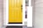 A front entryway with a bright yellow door featuring a black handle and lock. To the right, there is a narrow windowpane through which a small dog is peering outside. The siding of the house is light gray with a darker gray welcome mat on the concrete step.