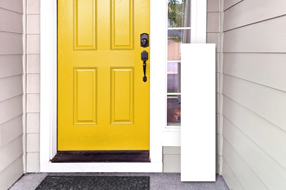 A front entryway with a bright yellow door featuring a black handle and lock. To the right, there is a narrow windowpane through which a small dog is peering outside. The siding of the house is light gray with a darker gray welcome mat on the concrete step.