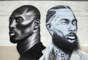 Two stylized monochrome portraits are painted side by side on a textured wall. The portrait on the left features a person with a shaved head and focused expression, rendered in dark gray and black tones. The portrait on the right shows a person with detailed braided hair, facial hair, and tattoos with inscriptions like 'PROLIFIC' and 'GOD WILL RISE'. Both portraits convey a strong sense of depth and realism, with shadows and highlights enhancing the features.