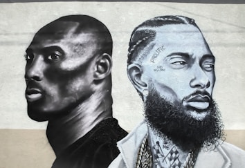 Two stylized monochrome portraits are painted side by side on a textured wall. The portrait on the left features a person with a shaved head and focused expression, rendered in dark gray and black tones. The portrait on the right shows a person with detailed braided hair, facial hair, and tattoos with inscriptions like 'PROLIFIC' and 'GOD WILL RISE'. Both portraits convey a strong sense of depth and realism, with shadows and highlights enhancing the features.