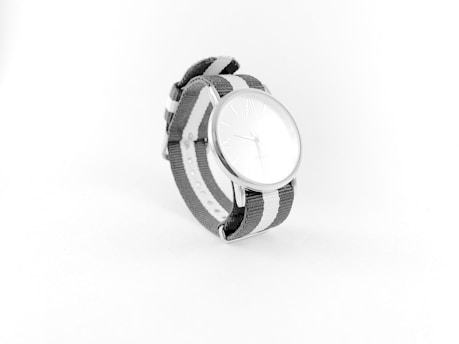A minimalist wristwatch with a sleek, silver round face and an analog display is centered in the image. The watch features a fabric strap in gray and white stripes, creating a modern and stylish appearance. The background remains plain and light, enhancing the watch's clean design.
