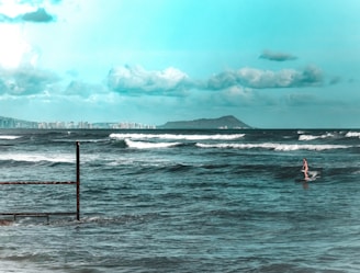 A surfer rides a wave in the foreground, with the ocean stretching out toward the horizon. In the distance, a city skyline and a mountain are visible under a sky filled with scattered clouds. The waves are gently crashing against a partially submerged metal structure.