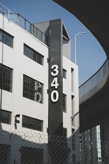 A modern, white building with large windows and a prominent black vertical sign featuring the number '340' in white font. There is an overpass or elevated road curving around the building, a chain-link fence in the foreground, and a light pole extending above the building. The sky is clear and blue, suggesting good weather.
