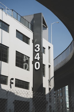 A modern, white building with large windows and a prominent black vertical sign featuring the number '340' in white font. There is an overpass or elevated road curving around the building, a chain-link fence in the foreground, and a light pole extending above the building. The sky is clear and blue, suggesting good weather.