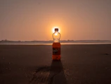 The orange élánmag bottle standing on a beach towel with a smartphone magnetically attached, waves in background.