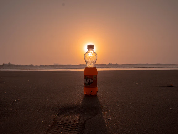 The orange élánmag bottle standing on a beach towel with a smartphone magnetically attached, waves in background.