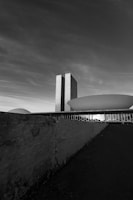 A black and white image captures a modern architectural scene featuring a tall, rectangular building with a textured facade and adjacent structures with dome-like shapes. The foreground includes a rough, angled wall leading towards the buildings, creating a strong sense of perspective under a cloudy sky.
