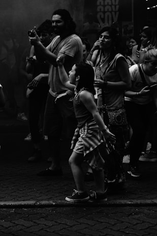 A group of people stands closely together, with some holding cameras or phones. The atmosphere appears to be lively, with a child in the foreground gesturing animatedly. The image is in black and white, giving it a classic or timeless feel.