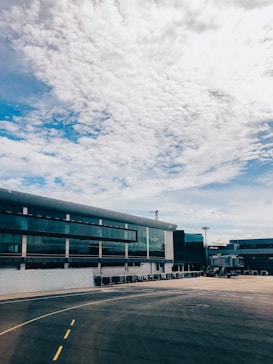 A modern airport terminal building with large glass windows, adjacent to an empty tarmac. The sky above is partly cloudy with a mix of white clouds and patches of blue sky. There are several airport ground service vehicles and equipment lined up near the terminal.