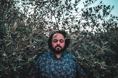 A person with closed eyes stands surrounded by dense foliage, giving a sense of tranquility and immersion in nature. The background consists of leafy branches, and the person's expression appears peaceful and contemplative.