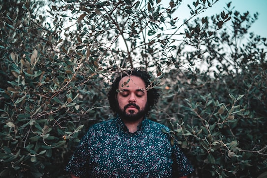 A person with closed eyes stands surrounded by dense foliage, giving a sense of tranquility and immersion in nature. The background consists of leafy branches, and the person's expression appears peaceful and contemplative.