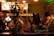 A group of people socializing in a dimly lit bar, with one individual drinking and another engaged in conversation. The scene is viewed through a window with gold lettering that reads 'Stock Brokers.' A menu with drink options is visible in the background.