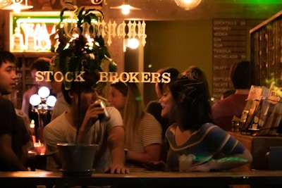 A group of people socializing in a dimly lit bar, with one individual drinking and another engaged in conversation. The scene is viewed through a window with gold lettering that reads 'Stock Brokers.' A menu with drink options is visible in the background.