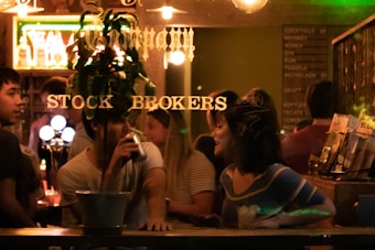 A group of people socializing in a dimly lit bar, with one individual drinking and another engaged in conversation. The scene is viewed through a window with gold lettering that reads 'Stock Brokers.' A menu with drink options is visible in the background.
