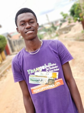 A person is wearing a purple t-shirt with branding related to a pharmaceutical product standing outdoors on a pathway. The background shows a blurred view of a residential area with greenery and structures.