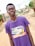 A person is wearing a purple t-shirt with branding related to a pharmaceutical product standing outdoors on a pathway. The background shows a blurred view of a residential area with greenery and structures.
