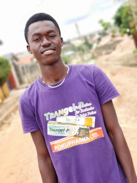 A person is wearing a purple t-shirt with branding related to a pharmaceutical product standing outdoors on a pathway. The background shows a blurred view of a residential area with greenery and structures.