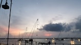 A peaceful waterfront scene during sunset with several large Chinese fishing nets silhouetted against the colorful sky. The water reflects the warm colors of the sunset, and there are people in the distance near the nets.