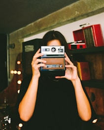 A person holds an old-fashioned Kodak instant camera in front of their face, positioned in a room with ambient lighting and a vintage setup. Shelves with retro items are visible in the background.
