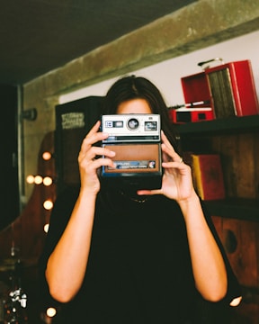 A person holds an old-fashioned Kodak instant camera in front of their face, positioned in a room with ambient lighting and a vintage setup. Shelves with retro items are visible in the background.