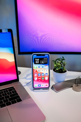 An office desk setup with a laptop, a large monitor displaying a colorful abstract wallpaper, and a smartphone on a stand with a calendar and app icons visible. A small potted plant adds a touch of greenery beside a sleek, metallic object holder.