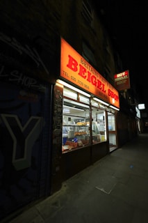 A brightly lit neon sign advertises a bagel shop, claiming to be Britain's first and best, established in 1855. The shop's interior is visible through large windows, showing bakery items like bagels on display. The surrounding area appears dimly lit with graffiti on the adjacent wall.