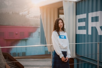 A person is standing in front of a backdrop of colorful shipping containers. The sunlight creates a soft glow, and the setting appears urban with industrial elements. The person is wearing a white sweater with a blue logo.