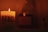 A lit candle with 'REBEL' written on it casts a warm glow in a dimly lit setting. To the side, there is a small bottle, and in the background, blurred objects add depth to the scene.