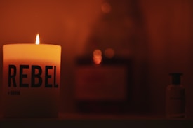 A lit candle with 'REBEL' written on it casts a warm glow in a dimly lit setting. To the side, there is a small bottle, and in the background, blurred objects add depth to the scene.