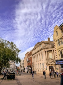 A street scene featuring a historic building with ornate architecture labeled Lloyds Bank. The sky is covered with a layer of textured clouds. People are walking along the street, and a few bicycles are parked near the building. The scene includes a mix of trees and other storefronts, giving an urban setting.