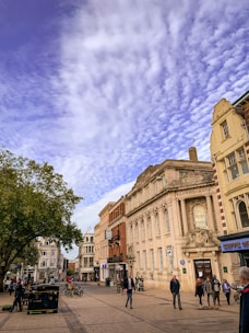 A street scene featuring a historic building with ornate architecture labeled Lloyds Bank. The sky is covered with a layer of textured clouds. People are walking along the street, and a few bicycles are parked near the building. The scene includes a mix of trees and other storefronts, giving an urban setting.