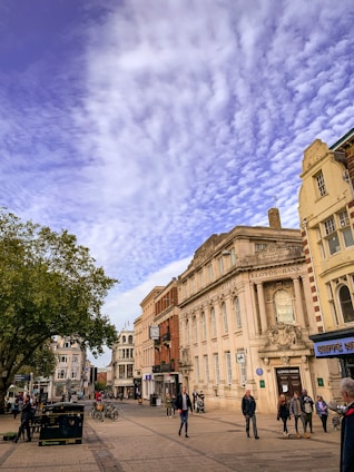 A street scene featuring a historic building with ornate architecture labeled Lloyds Bank. The sky is covered with a layer of textured clouds. People are walking along the street, and a few bicycles are parked near the building. The scene includes a mix of trees and other storefronts, giving an urban setting.